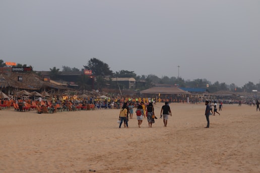 A sandy beach with several people walking along the shore, some in groups and others alone. On the left side, there are brightly colored beach chairs and huts with signage indicating restaurants or bars. The sky is overcast, adding a muted tone to the scene. In the background, trees and additional structures are visible.