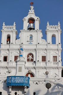 A white colonial-style building with arched windows and detailed architectural features, topped with a cross. There's a prominent bell and a statue of a religious figure on a pedestal. The structure includes a blue balcony railing and several spherical lamps.