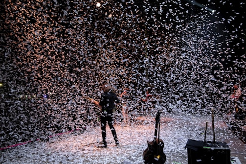 A musician stands on a stage surrounded by a flurry of confetti falling from above. The background is dark, with stage lights partially illuminating the scene, creating a dynamic and festive atmosphere. Several musical instruments and equipment are visible on the stage.