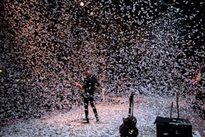 A musician stands on a stage surrounded by a flurry of confetti falling from above. The background is dark, with stage lights partially illuminating the scene, creating a dynamic and festive atmosphere. Several musical instruments and equipment are visible on the stage.