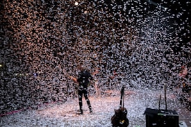 A musician stands on a stage surrounded by a flurry of confetti falling from above. The background is dark, with stage lights partially illuminating the scene, creating a dynamic and festive atmosphere. Several musical instruments and equipment are visible on the stage.
