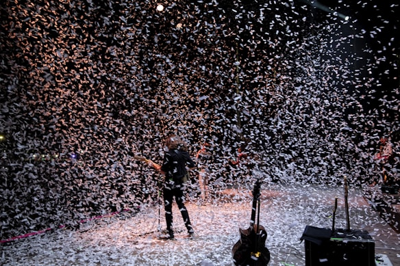 A musician stands on a stage surrounded by a flurry of confetti falling from above. The background is dark, with stage lights partially illuminating the scene, creating a dynamic and festive atmosphere. Several musical instruments and equipment are visible on the stage.