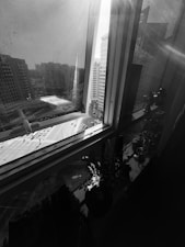 A cityscape view through a window with sunlight streaming in. The window has a partially open curtain, revealing tall buildings and a street below with visible cars. Inside, there are decorative items and a plant on the windowsill, casting shadows.