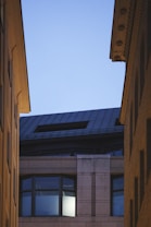 An urban scene features the upper portion of a modern building, framed by the rooftops of two other structures converging towards the sky. The building has large windows, one of which is illuminated from within, suggesting evening or early morning. The architectural style includes clean lines and a mix of materials.
