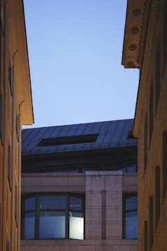 An urban scene features the upper portion of a modern building, framed by the rooftops of two other structures converging towards the sky. The building has large windows, one of which is illuminated from within, suggesting evening or early morning. The architectural style includes clean lines and a mix of materials.