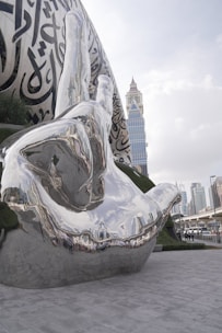 A large, reflective metallic sculpture of a hand is prominently displayed in an urban setting, featuring intricate Arabic calligraphy on its surface. The background includes a modern city skyline with tall buildings and overcast skies.