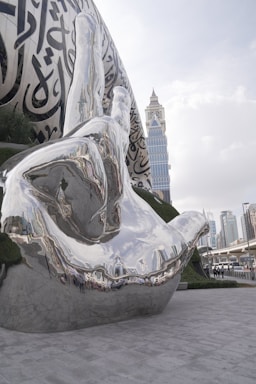 A large, reflective metallic sculpture of a hand is prominently displayed in an urban setting, featuring intricate Arabic calligraphy on its surface. The background includes a modern city skyline with tall buildings and overcast skies.