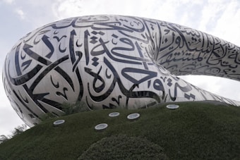 A futuristic architectural structure with flowing curves and adorned with large, stylized Arabic calligraphy. The building's exterior has a sleek metallic finish, set against a backdrop of a partially cloudy sky. The foreground features lush green landscaping with round skylights.