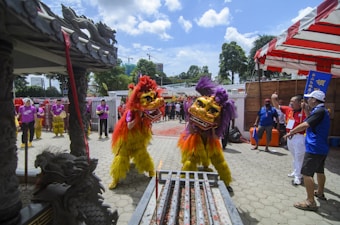 Two performers in bright yellow and red traditional lion dance costumes engage in a dynamic lion dance. Their costumes feature golden masks and vibrant feather-like details. Surrounding them are people in purple shirts, possibly part of a musical ensemble, along with others watching the performance. The setting has traditional Asian architectural elements and festive decorations, suggesting a cultural celebration outdoors under a clear blue sky.