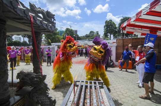 Two performers in bright yellow and red traditional lion dance costumes engage in a dynamic lion dance. Their costumes feature golden masks and vibrant feather-like details. Surrounding them are people in purple shirts, possibly part of a musical ensemble, along with others watching the performance. The setting has traditional Asian architectural elements and festive decorations, suggesting a cultural celebration outdoors under a clear blue sky.