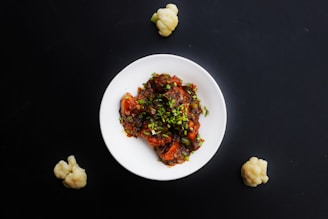 A white plate is filled with a colorful dish consisting of what appears to be chicken or tofu pieces in a rich, dark sauce garnished with chopped green onions. The background is a deep black surface, and three small cauliflower florets are placed around the plate.