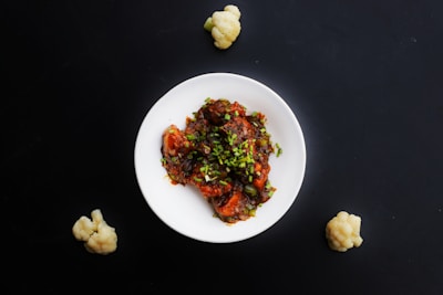 A white plate is filled with a colorful dish consisting of what appears to be chicken or tofu pieces in a rich, dark sauce garnished with chopped green onions. The background is a deep black surface, and three small cauliflower florets are placed around the plate.