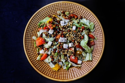A vibrant salad is arranged in a textured circular dish against a dark background. The salad contains mixed greens, cut tomatoes, cubes of cheese, various seeds, bell peppers, and small legumes, combining fresh and colorful ingredients for a visually appealing dish.