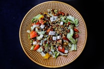 A vibrant salad is arranged in a textured circular dish against a dark background. The salad contains mixed greens, cut tomatoes, cubes of cheese, various seeds, bell peppers, and small legumes, combining fresh and colorful ingredients for a visually appealing dish.