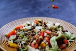 A fresh salad featuring an assortment of colorful vegetables, including chopped tomatoes, cucumbers, and bell peppers. There are leaves of lettuce and cubes of white cheese scattered throughout. Crunchy seeds and nuts are sprinkled on top, adding texture and flavor.