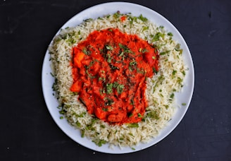A white plate contains a serving of basmati rice garnished with green herbs, surrounding a vibrant red curry with visible spices and herbs sprinkled on top. The dish is set on a dark surface.