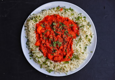 A white plate contains a serving of basmati rice garnished with green herbs, surrounding a vibrant red curry with visible spices and herbs sprinkled on top. The dish is set on a dark surface.