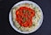 A white plate contains a serving of basmati rice garnished with green herbs, surrounding a vibrant red curry with visible spices and herbs sprinkled on top. The dish is set on a dark surface.