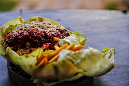 A close-up view of a dish with a base of leafy greens, topped with a generous serving of a chunky red sauce, possibly marinara or salsa, sprinkled with cheese. There are visible pieces of yellow and orange vegetables on the side.