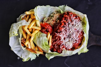 A dish served in a cabbage leaf featuring a portion of crispy French fries on the left and a mixture of rice topped with a rich, red chili sauce and melted cheese on the right. There is a roasted tomato situated between the two main components.