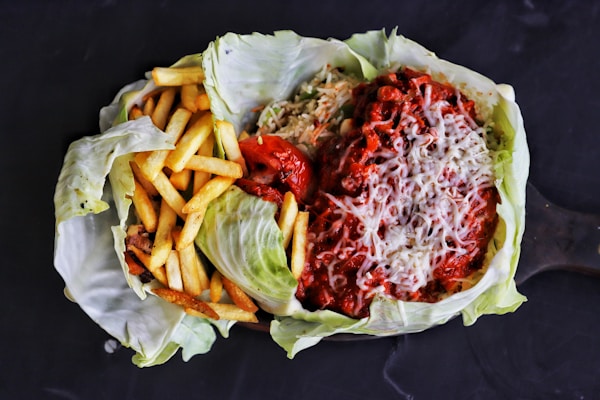 A dish served in a cabbage leaf featuring a portion of crispy French fries on the left and a mixture of rice topped with a rich, red chili sauce and melted cheese on the right. There is a roasted tomato situated between the two main components.