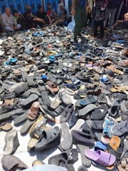 A large pile of various shoes and sandals is spread on the ground, surrounded by people who appear to be seated or standing nearby. The footwear is predominantly dark, with occasional spots of bright colors. The background features several people, some sitting against a wall.