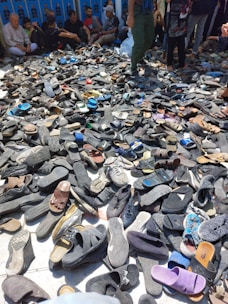 A large pile of various shoes and sandals is spread on the ground, surrounded by people who appear to be seated or standing nearby. The footwear is predominantly dark, with occasional spots of bright colors. The background features several people, some sitting against a wall.