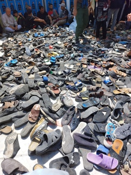 A large pile of various shoes and sandals is spread on the ground, surrounded by people who appear to be seated or standing nearby. The footwear is predominantly dark, with occasional spots of bright colors. The background features several people, some sitting against a wall.