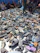 A large pile of various shoes and sandals is spread on the ground, surrounded by people who appear to be seated or standing nearby. The footwear is predominantly dark, with occasional spots of bright colors. The background features several people, some sitting against a wall.