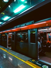 A metro train stationed at a platform with its doors open, allowing passengers to board or alight. The interior lights of the train and the station create a warm and vibrant ambiance. People are visible inside the train, some seated, while others stand near the doors. The platform has clear safety markings and instructions written in multiple languages.