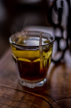 A steaming glass of tea sits on a wooden table, with the warmth visible through the rising steam. The tea has a rich amber color, and a metal spoon rests inside the glass, catching the light. The background is softly blurred, adding to the cozy, inviting atmosphere.