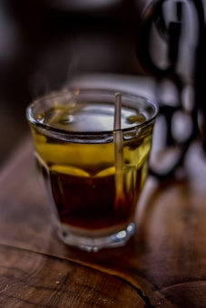 A steaming glass of tea sits on a wooden table, with the warmth visible through the rising steam. The tea has a rich amber color, and a metal spoon rests inside the glass, catching the light. The background is softly blurred, adding to the cozy, inviting atmosphere.