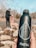 A close-up view of a dark reusable water bottle with water droplets on its surface, held in a person's hand. In the background, a person dressed in dark clothing is walking a dog on a leash by a rocky area near water. The scene is set outdoors, with trees visible without leaves, suggesting an autumn or winter setting.