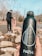 A close-up view of a dark reusable water bottle with water droplets on its surface, held in a person's hand. In the background, a person dressed in dark clothing is walking a dog on a leash by a rocky area near water. The scene is set outdoors, with trees visible without leaves, suggesting an autumn or winter setting.
