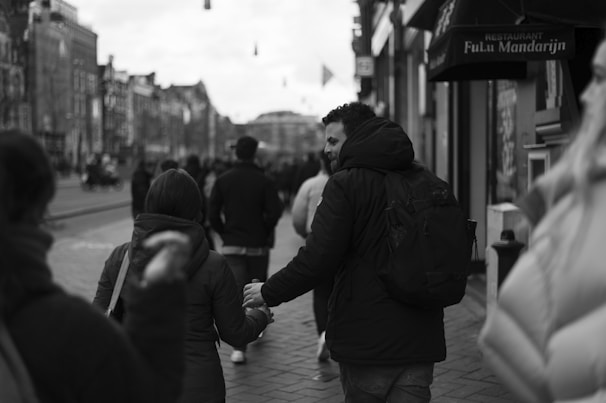 A black and white photo depicting people walking down a city street. A man with a backpack holds hands with another person, suggesting a sense of connection. Urban buildings and storefronts line the street, and other pedestrians are visible in the background.