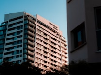 A modern high-rise apartment building under a clear blue sky. The structure features numerous balconies and large windows, with reflective surfaces. The image captures a section of the urban environment, where another building partially frames the view.