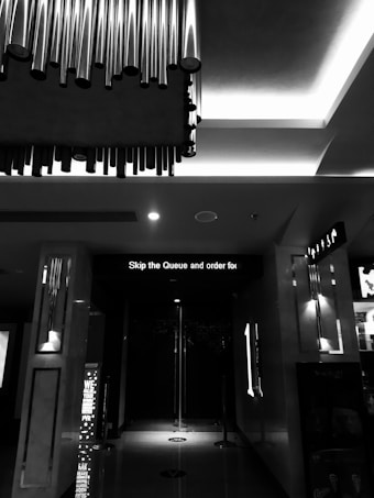 A monochrome image of an indoor setting with modern ceiling lights and metallic pipe decorations. The floor reflects some light, and there's a sign displaying 'Skip the Queue and order' above an entrance. The surroundings appear sleek and minimalist.