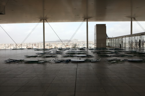 A spacious modern terrace with sleek, minimal design overlooking a cityscape. The area is covered and supported by large columns and cables, with numerous green lounge chairs arranged flatly on the tiled floor. A glass barrier runs along the edge, and a building structure is visible in the background.