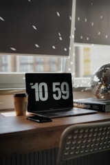 A cozy workspace with a laptop displaying the time 10:59. Sunlight filters through window blinds, casting dotted light patterns on the wall. A disposable coffee cup is placed next to the laptop, accompanied by a smartphone. A photography book and a decorative disco ball add aesthetic elements to the wooden desk.