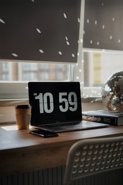 A cozy workspace with a laptop displaying the time 10:59. Sunlight filters through window blinds, casting dotted light patterns on the wall. A disposable coffee cup is placed next to the laptop, accompanied by a smartphone. A photography book and a decorative disco ball add aesthetic elements to the wooden desk.