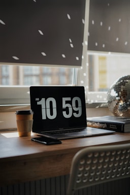 A cozy workspace with a laptop displaying the time 10:59. Sunlight filters through window blinds, casting dotted light patterns on the wall. A disposable coffee cup is placed next to the laptop, accompanied by a smartphone. A photography book and a decorative disco ball add aesthetic elements to the wooden desk.