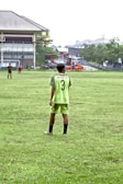 A person stands on a grassy soccer field wearing a neon green sports jersey with the number 3 and 'Almer FC' written on the back. In the background, there are other players, a large building, trees, and some urban structures.