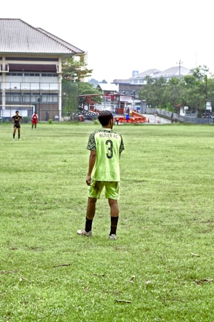 A person stands on a grassy soccer field wearing a neon green sports jersey with the number 3 and 'Almer FC' written on the back. In the background, there are other players, a large building, trees, and some urban structures.