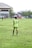A person stands on a grassy soccer field wearing a neon green sports jersey with the number 3 and 'Almer FC' written on the back. In the background, there are other players, a large building, trees, and some urban structures.