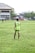 A person stands on a grassy soccer field wearing a neon green sports jersey with the number 3 and 'Almer FC' written on the back. In the background, there are other players, a large building, trees, and some urban structures.