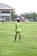 A person stands on a grassy soccer field wearing a neon green sports jersey with the number 3 and 'Almer FC' written on the back. In the background, there are other players, a large building, trees, and some urban structures.
