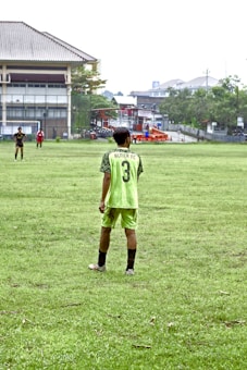 A person stands on a grassy soccer field wearing a neon green sports jersey with the number 3 and 'Almer FC' written on the back. In the background, there are other players, a large building, trees, and some urban structures.