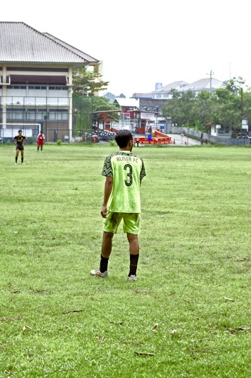 A person stands on a grassy soccer field wearing a neon green sports jersey with the number 3 and 'Almer FC' written on the back. In the background, there are other players, a large building, trees, and some urban structures.