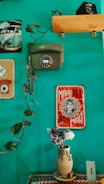 A teal-colored wall is decorated with vintage items, including a rotary dial telephone and a classic poster that reads 'Make Art Not War.' A vine with lush green leaves extends across the wall. Below, a crocheted tablecloth in various colors adorns a surface holding a rustic clay vase filled with a small bouquet of dried flowers. A postcard or card labeled with the number '4' is placed nearby, and a small photographic print of a vintage Volkswagen van is seen in the corner.