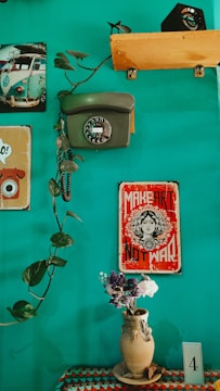 A teal-colored wall is decorated with vintage items, including a rotary dial telephone and a classic poster that reads 'Make Art Not War.' A vine with lush green leaves extends across the wall. Below, a crocheted tablecloth in various colors adorns a surface holding a rustic clay vase filled with a small bouquet of dried flowers. A postcard or card labeled with the number '4' is placed nearby, and a small photographic print of a vintage Volkswagen van is seen in the corner.